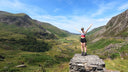 A woman stands on a stone wall in a mountain valley with her arms raised, wearing a pink and white sports top, black shorts, and running shoes under a bright blue sky.