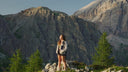 A woman stands on a rocky mountain ridge with pine trees and large rugged mountains in the background, wearing a light-colored jacket, dark shorts, and carrying a backpack.