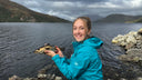 A smiling woman in a bright turquoise hooded jacket stands on rocky shores holding a fish, with a large lake and mountains in the background under a cloudy sky.