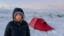 A woman wearing a navy blue winter puffer jacket with a beige hood lining stands in a snowy outdoor landscape next to a red dome tent.