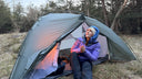 A person sitting inside the open doorway of a gray camping tent pitched in a grassy wooded outdoor setting, wearing a purple jacket and blue beanie.