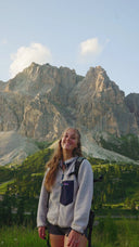 A smiling young woman stands outdoors in front of large rocky mountains, wearing a light gray fleece zip-up jacket with a dark pocket, dark shorts, and a backpack.