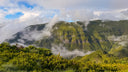 A scenic landscape of green mountains partially covered in white clouds and mist, with yellow-green shrubs in the foreground under a blue sky with clouds.
