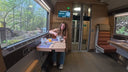 A woman sits at a small table inside a train carriage, preparing food. The train interior has wood-toned walls, a window showing a forest view, and a red upholstered seat nearby.
